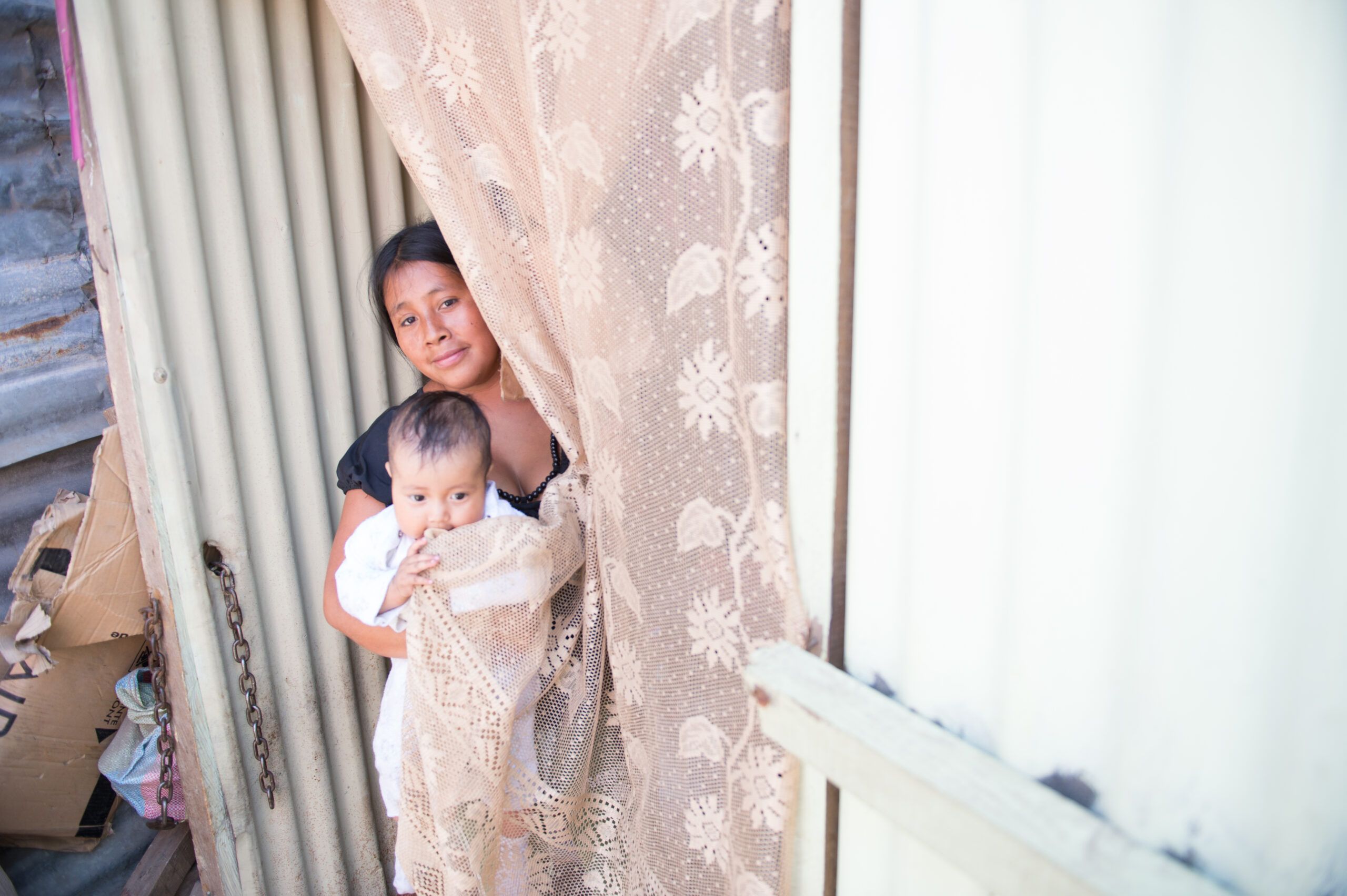 A woman holding a baby looks out from behind a curtain.