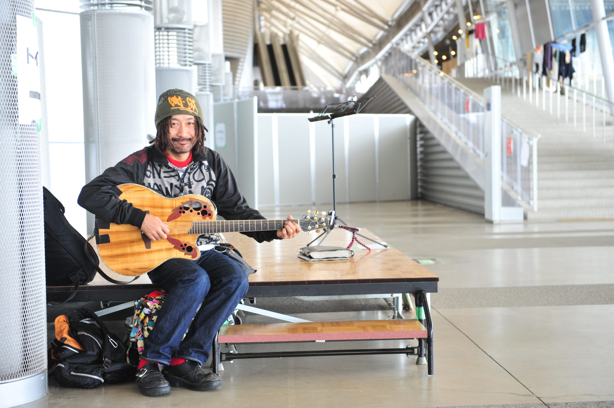 A Japanese man sitting on a stage playing guitar.