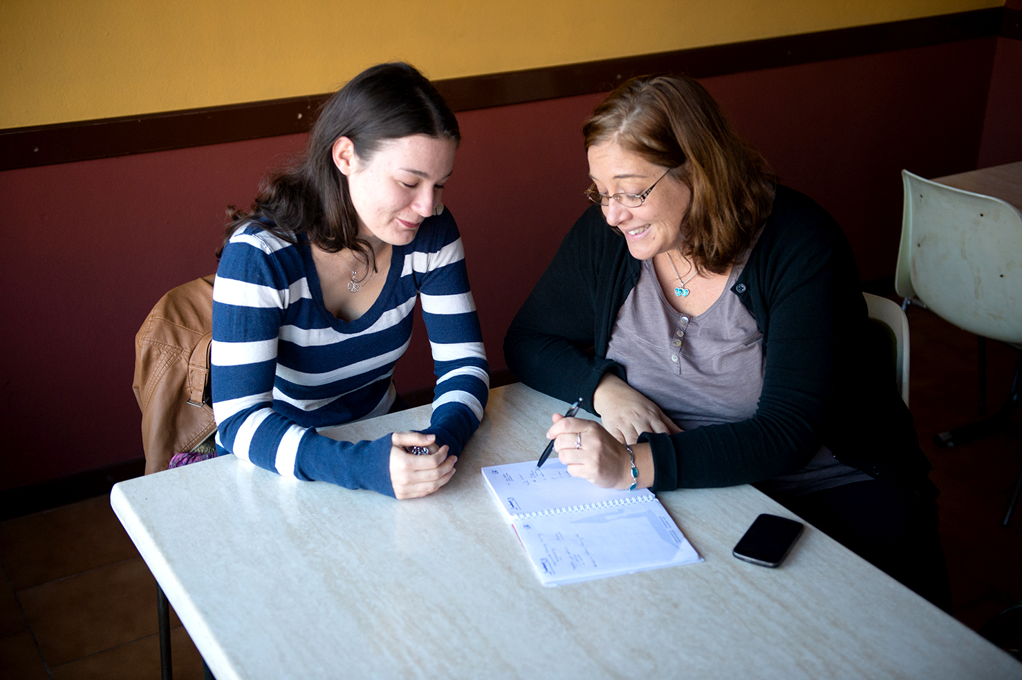 Two women sit at a table with a notebook talking.