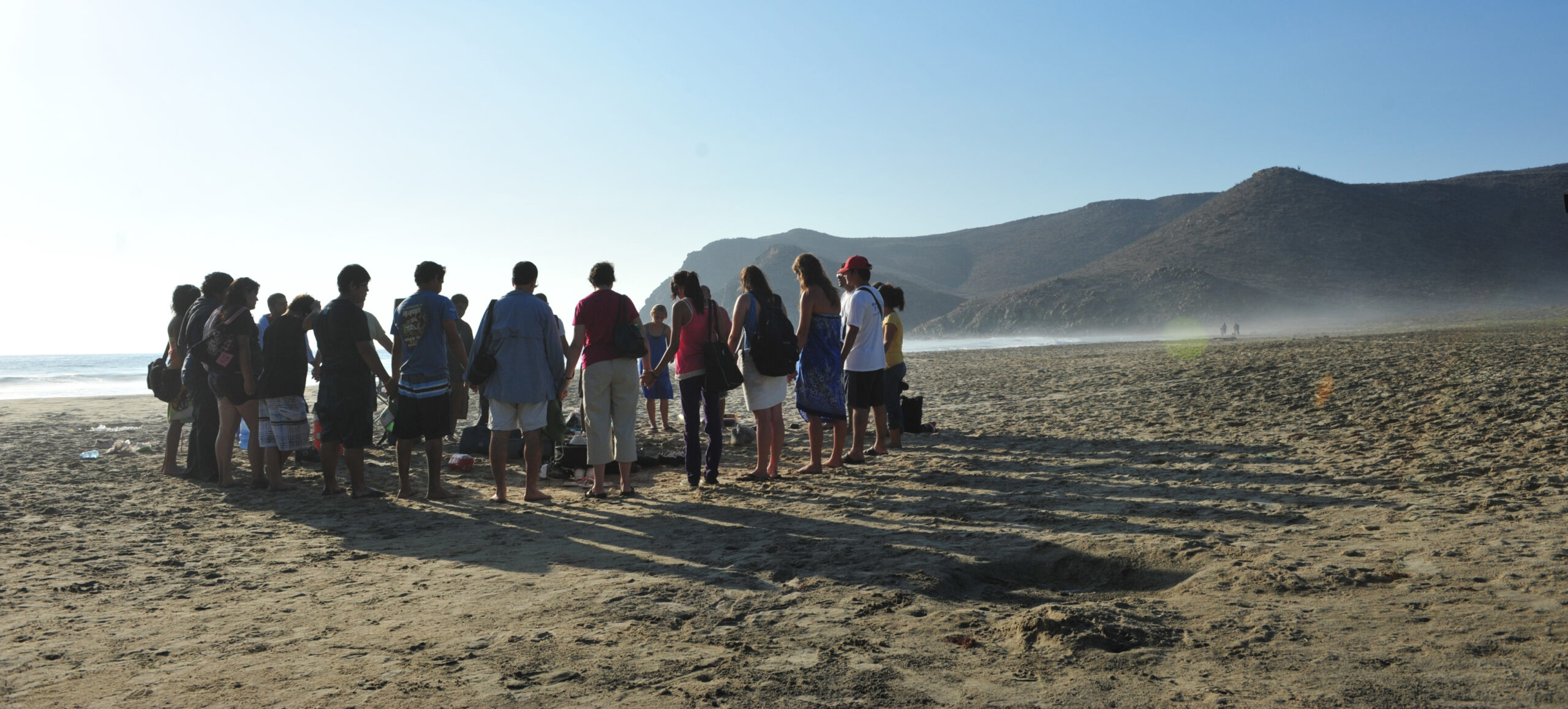 A group stands together praying while holding hands on a beach.