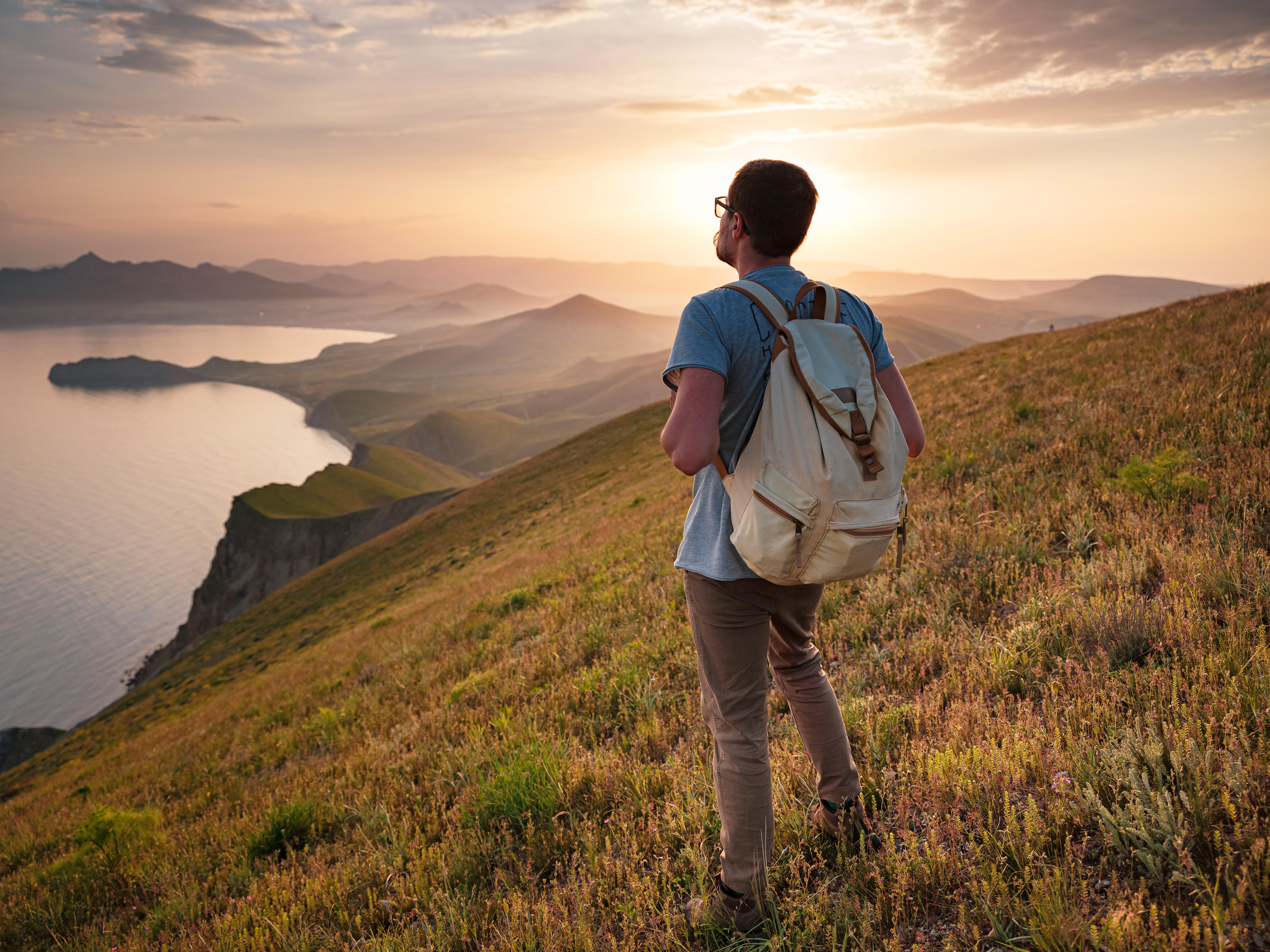 Young sporty man with backpack standing on the top of rock and looking at the seashore and mountains at sunset in summer. the lifestyle concept of traveling outdoors.