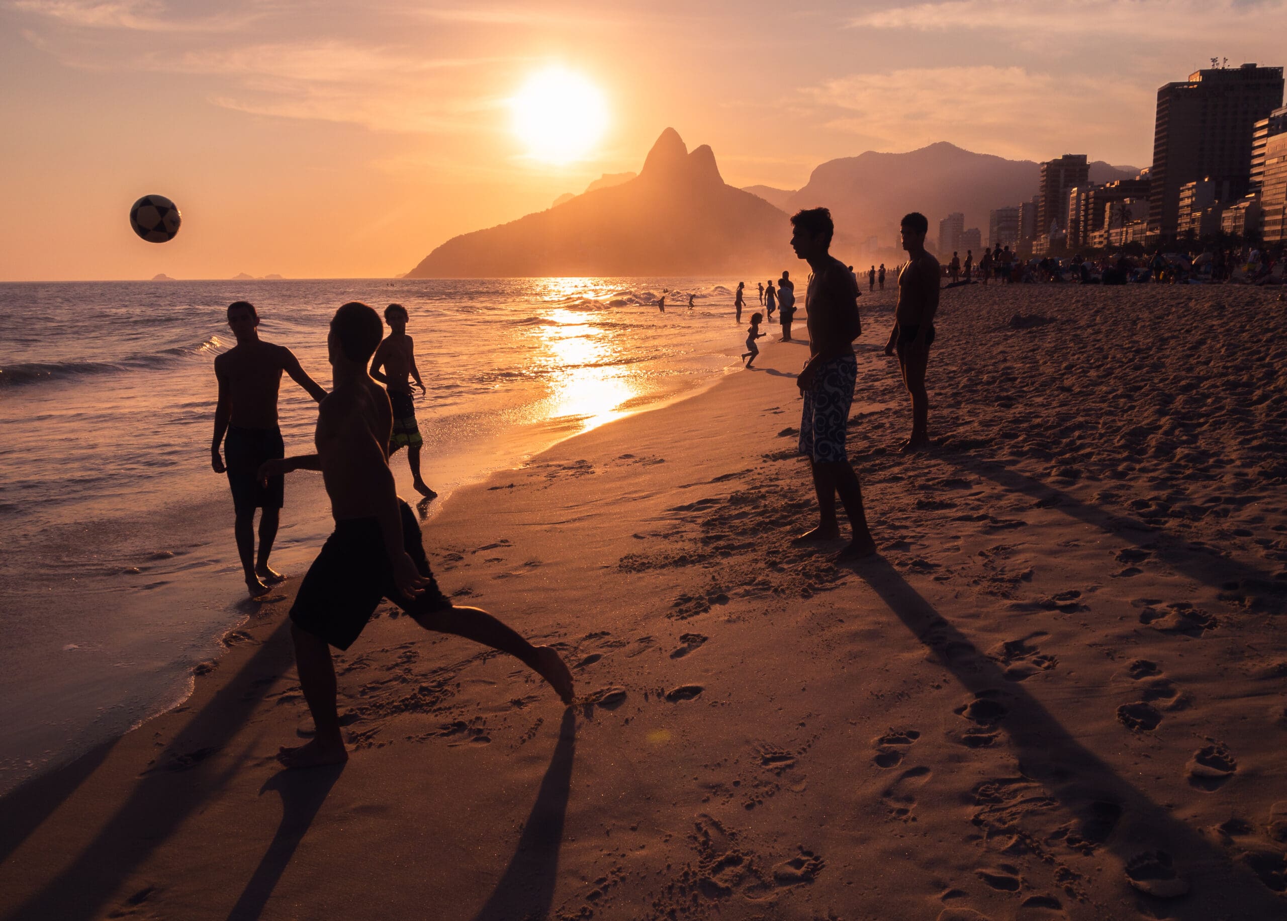 Teens playing soccer on the beach at sunset.